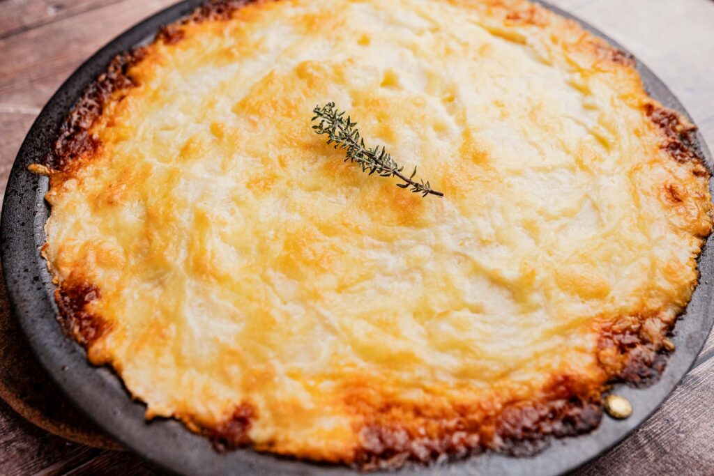 Baked vegetarian shepherd's pie topped with golden mashed potatoes and garnished with a sprig of thyme on a round black dish.