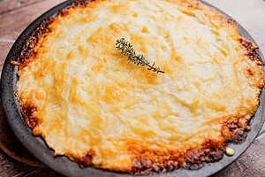 Baked vegetarian shepherd's pie topped with golden mashed potatoes and garnished with a sprig of thyme on a round black dish.