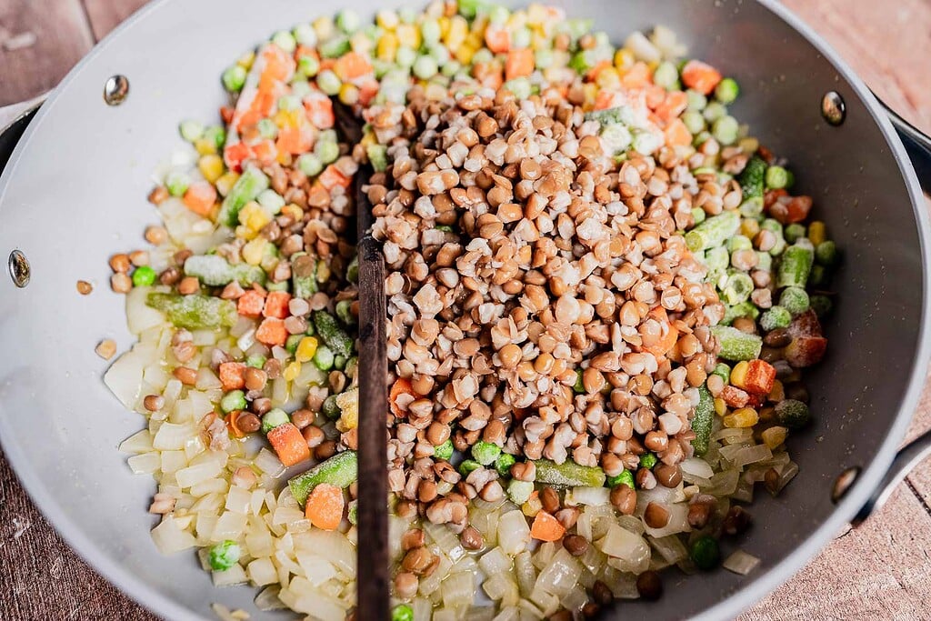 A pan contains cooked buckwheat, sautéed onions, and mixed vegetables—perfect as a hearty base for a vegetarian shepherd's pie, with a wooden spoon resting inside.