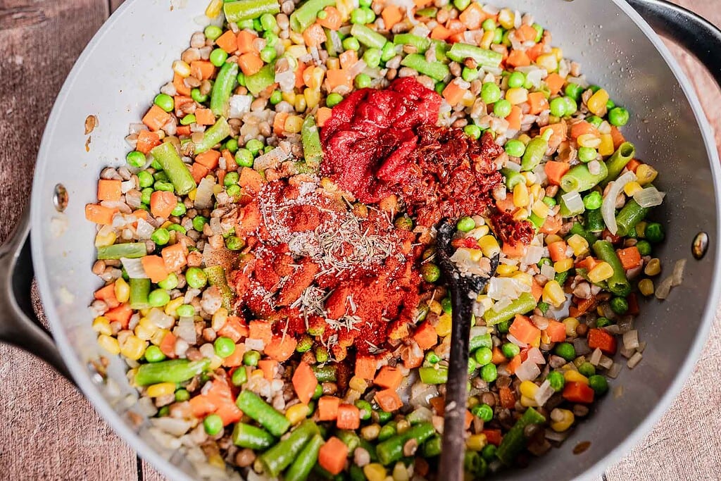 A skillet filled with mixed vegetables, peas, corn, green beans, carrots, tomato paste, and spices being sautéed with a spoon—perfect as a hearty base for vegetarian shepherd's pie.