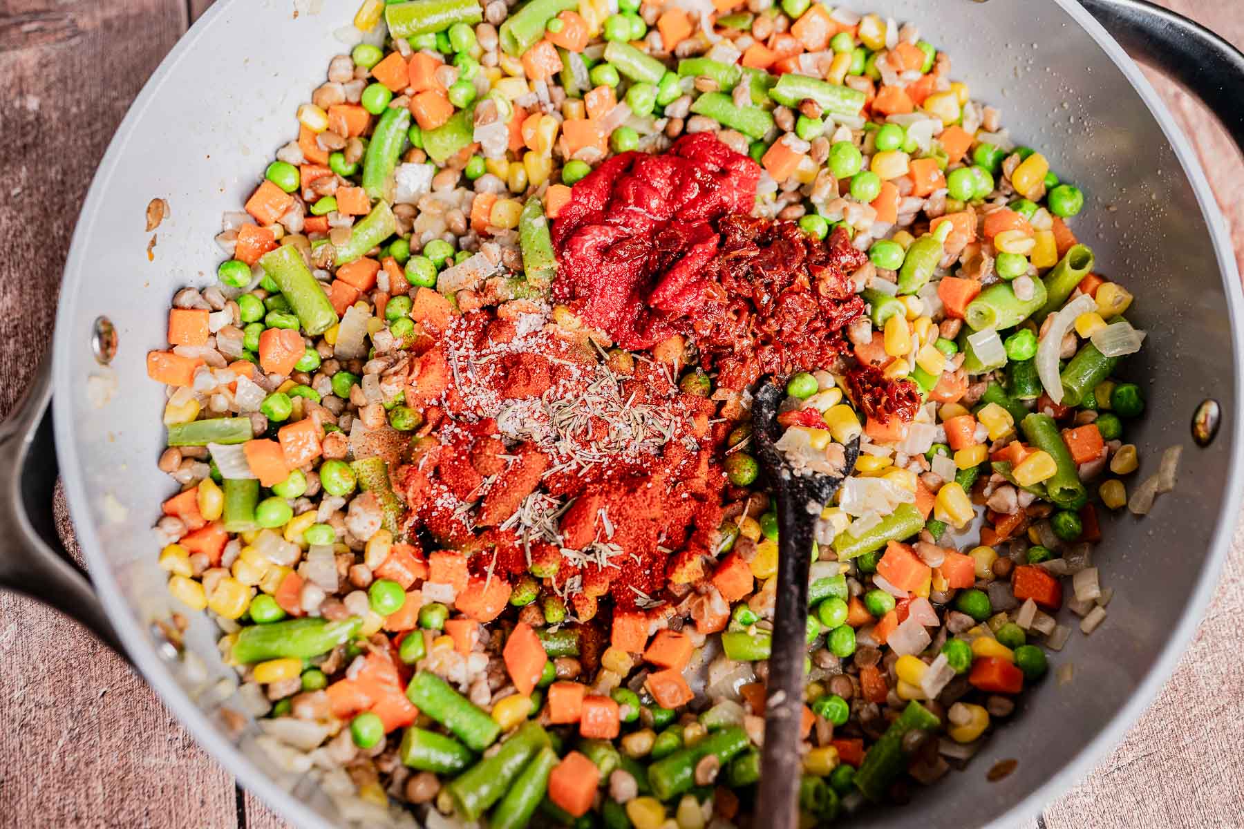 A skillet filled with mixed vegetables, peas, corn, green beans, carrots, tomato paste, and spices being sautéed with a spoon—perfect as a hearty base for vegetarian shepherd's pie.