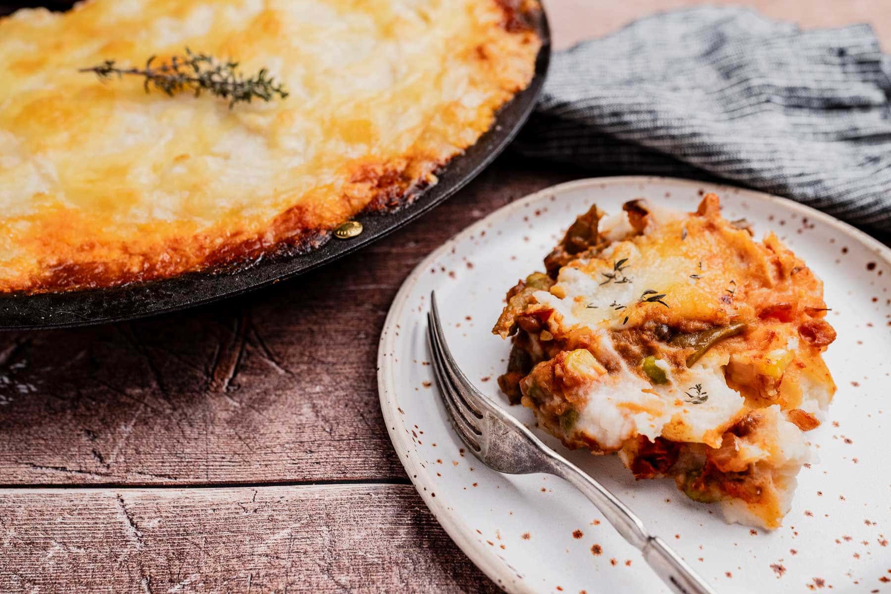 A slice of vegetable lasagna served on a speckled plate with a fork, next to the remaining lasagna in a baking dish—perfect alongside classics like vegetarian shepherd's pie.