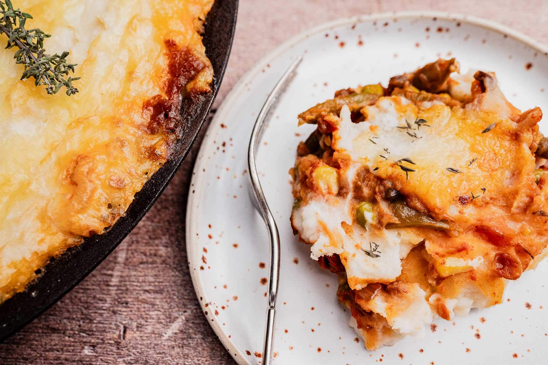 A serving of cheesy vegetarian shepherd's pie casserole with vegetables sits on a white plate beside a cast iron dish holding the remaining casserole.