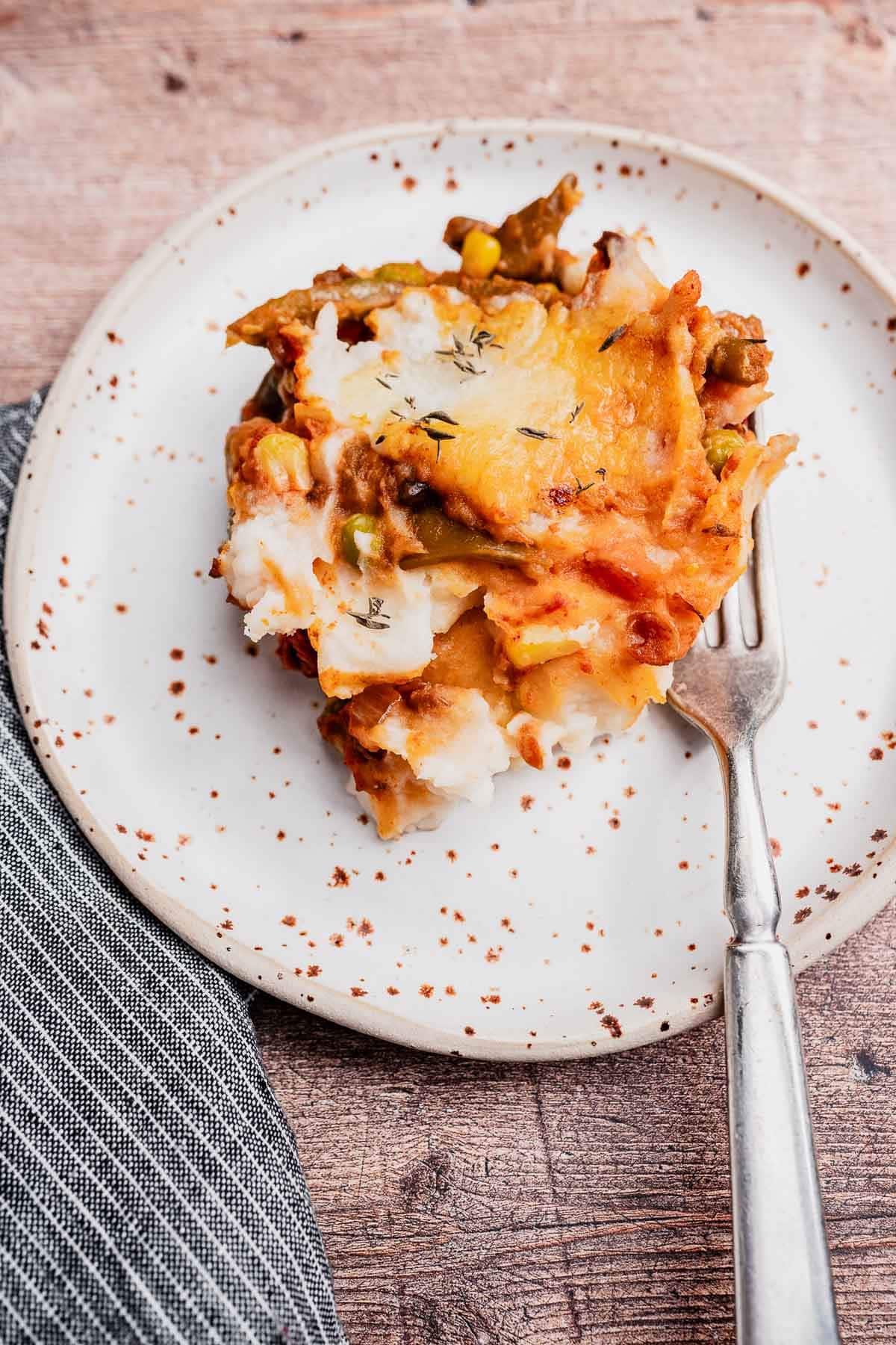A slice of vegetarian shepherd's pie with mashed potatoes and vegetables sits on a speckled white plate, with a fork beside it on a wooden table.