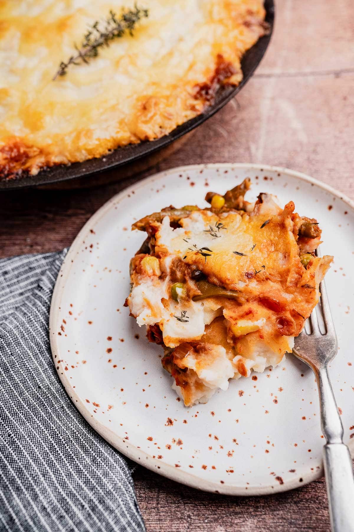 A plated serving of vegetarian shepherd's pie with mashed potato topping on a white speckled plate and a fork, with the remainder of the pie in a pan in the background.