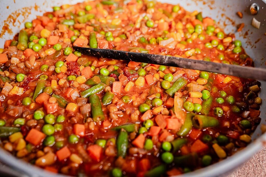 A close-up of a pan filled with mixed vegetables, including peas, carrots, green beans, and corn, in a tomato-based sauce—perfect as a filling for vegetarian shepherd's pie—with a black spatula resting inside.