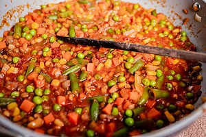 A close-up of a pan filled with mixed vegetables, including peas, carrots, green beans, and corn, in a tomato-based sauce—perfect as a filling for vegetarian shepherd's pie—with a black spatula resting inside.