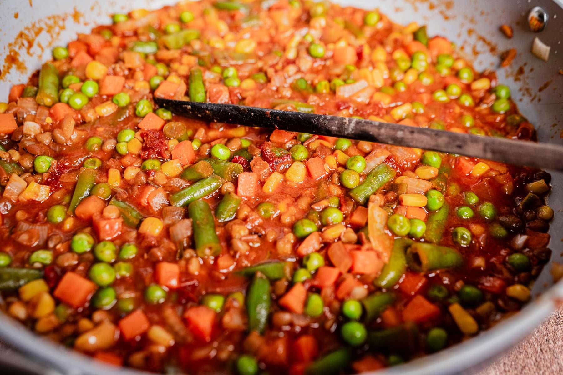 A close-up of a pan filled with mixed vegetables, including peas, carrots, green beans, and corn, in a tomato-based sauce—perfect as a filling for vegetarian shepherd's pie—with a black spatula resting inside.
