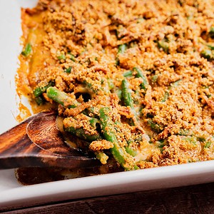 A wooden spoon serving gluten free vegan green bean casserole topped with crispy breadcrumbs from a white baking dish.