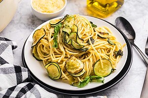 A plate of spaghetti topped with roasted zucchini slices and fresh basil, with a small bowl of grated cheese and a bottle of olive oil in the background.