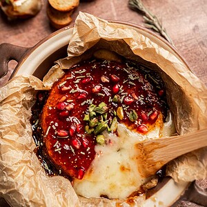 Baked brie and fig jam cheese topped with pomegranate seeds, pistachios, herbs, and sauce, served in parchment paper with a wooden spoon.
