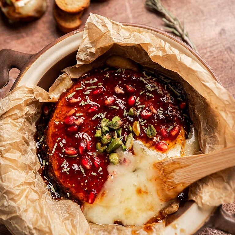 Baked brie and fig jam cheese topped with pomegranate seeds, pistachios, herbs, and sauce, served in parchment paper with a wooden spoon.