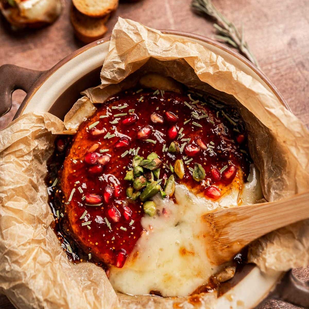 Baked brie and fig jam cheese topped with pomegranate seeds, pistachios, herbs, and sauce, served in parchment paper with a wooden spoon.