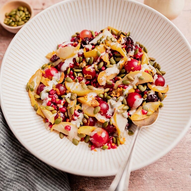 A bowl filled with a festive thanksgiving fruit salad of grapes, pear slices, pomegranate seeds, pumpkin seeds, dried cranberries, and drizzled with a creamy dressing. A spoon rests in the bowl.