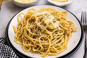 A plate of spaghetti pasta topped with grated cheese and black pepper, placed on a white plate with a fork nearby.