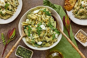 A bowl of pasta with pesto, arugula, ricotta, and pine nuts, surrounded by sliced bread, pesto sauce, pine nuts, and garnished with green leaves.