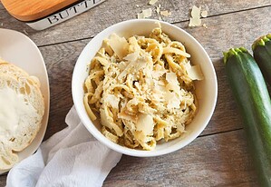 A bowl of pasta topped with shredded cheese sits on a wooden surface next to sliced bread with butter and two whole zucchinis.