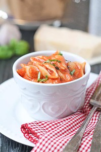 A white bowl of pasta with red tomato sauce, garnished with chopped herbs, sits on a white plate with a red-checked napkin and cutlery beside it.