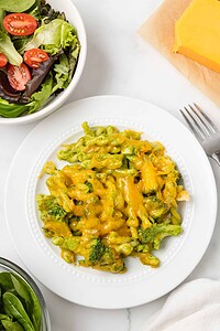 A plate of broccoli casserole topped with melted cheddar cheese sits next to a bowl of salad and a block of cheddar cheese—perfect inspiration for anyone seeking hearty vegetarian pasta recipes.