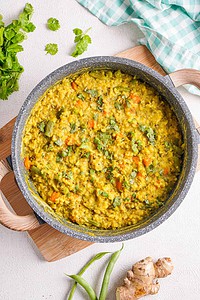 A pan of cooked khichdi, a yellow Indian rice and lentil dish with visible herbs and vegetables, sits on a wooden board with ginger, green chilies, and cilantro nearby.
