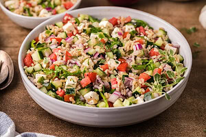 A white bowl filled with a salad containing chopped cucumber, tomato, red onion, herbs, rice, and cubes of feta cheese.