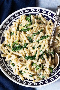 A plate of pasta with creamy white sauce and wilted spinach, served with a silver spoon on a patterned dish.