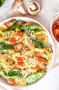 A plate of rotini pasta with spinach, mushrooms, cherry tomatoes, and grated cheese—perfect for those seeking delicious vegetarian pasta recipes—with fresh mushrooms and a bowl of tomatoes nearby.
