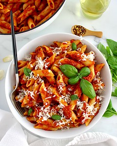 A bowl of penne pasta with tomato sauce, grated cheese, and basil leaves, with a fork and fresh basil, red pepper flakes, and olive oil on the side.