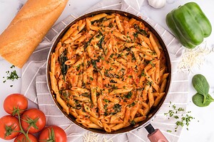 A skillet filled with penne pasta in a creamy tomato sauce, garnished with herbs, surrounded by bread, green bell pepper, tomatoes, and spinach.