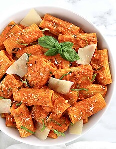 A bowl of rigatoni pasta with red tomato sauce, topped with parmesan cheese shavings and fresh basil leaves.