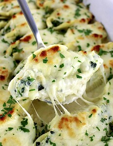 A close-up of a cheesy, baked stuffed pasta shell with spinach and ricotta, topped with melted cheese and chopped parsley, being lifted from a baking dish.