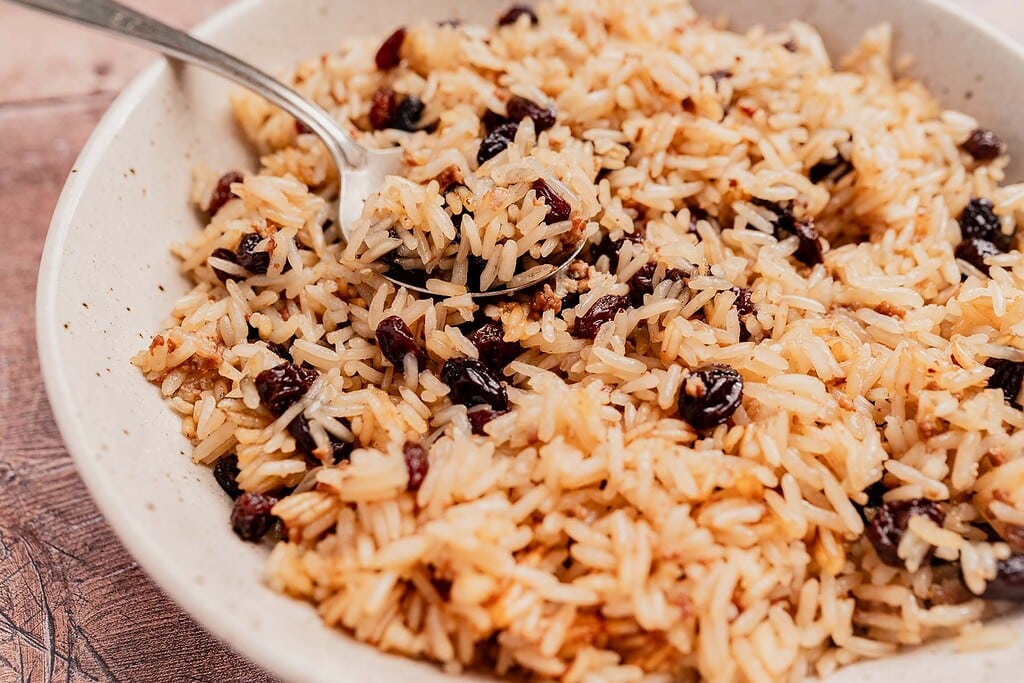 A bowl of cooked rice mixed with raisins is served with a spoon resting inside, perfect for those seeking delicious vegetarian rice recipes.
