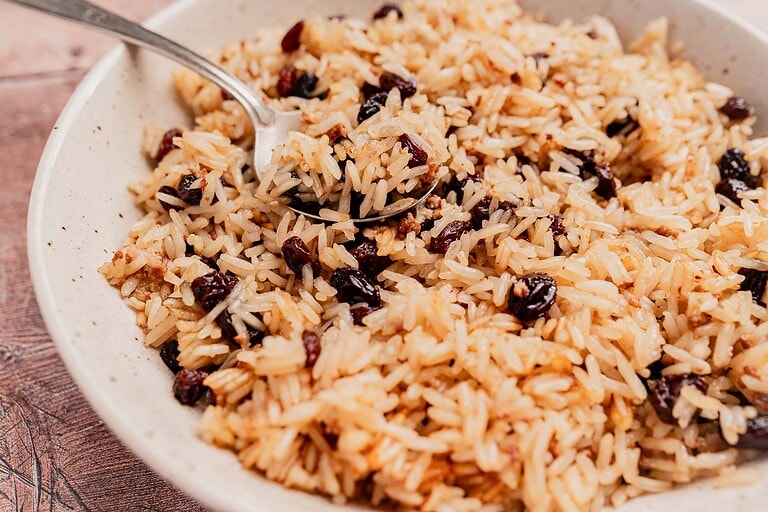 A bowl of cooked rice mixed with raisins is served with a spoon resting inside, perfect for those seeking delicious vegetarian rice recipes.