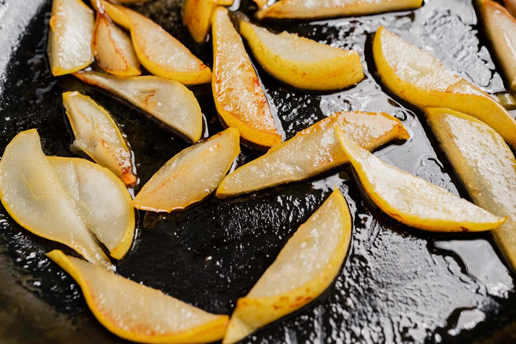 Sliced pears, perfect for a Thanksgiving fruit salad, being cooked and caramelized in oil on a black skillet.