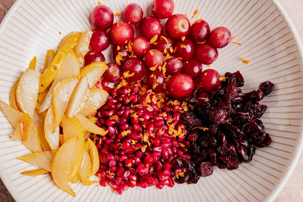 A plate with sections of sliced pears, red grapes, pomegranate seeds, and dried cranberries, garnished with orange zest—a delightful Thanksgiving fruit salad.