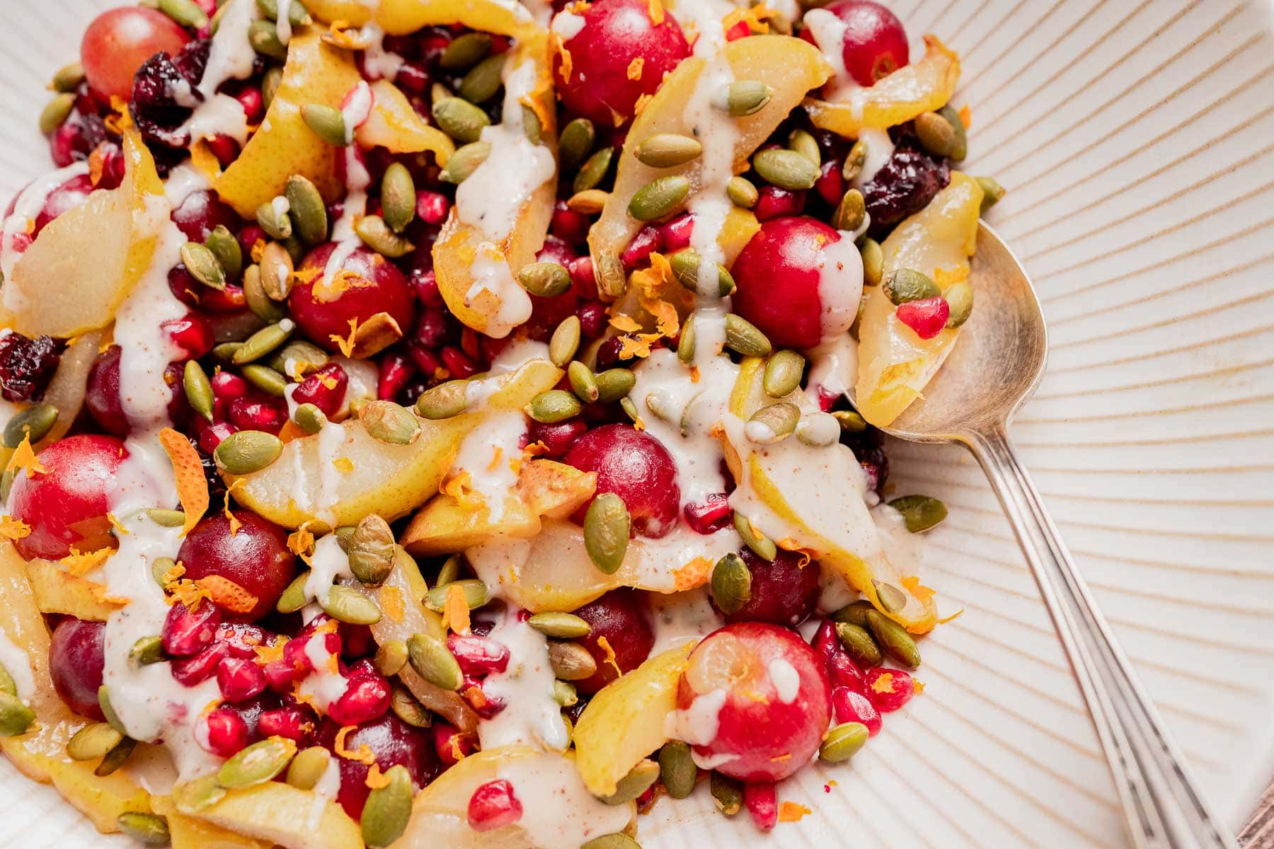 A bowl of Thanksgiving fruit salad with grapes, sliced pears, pomegranate seeds, pepitas, and a creamy drizzle, served with a spoon.