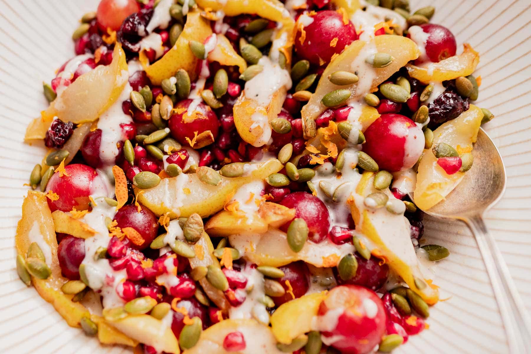 A bowl of thanksgiving fruit salad with sliced pears, red grapes, pomegranate seeds, pumpkin seeds, and a creamy drizzle, served with a spoon.