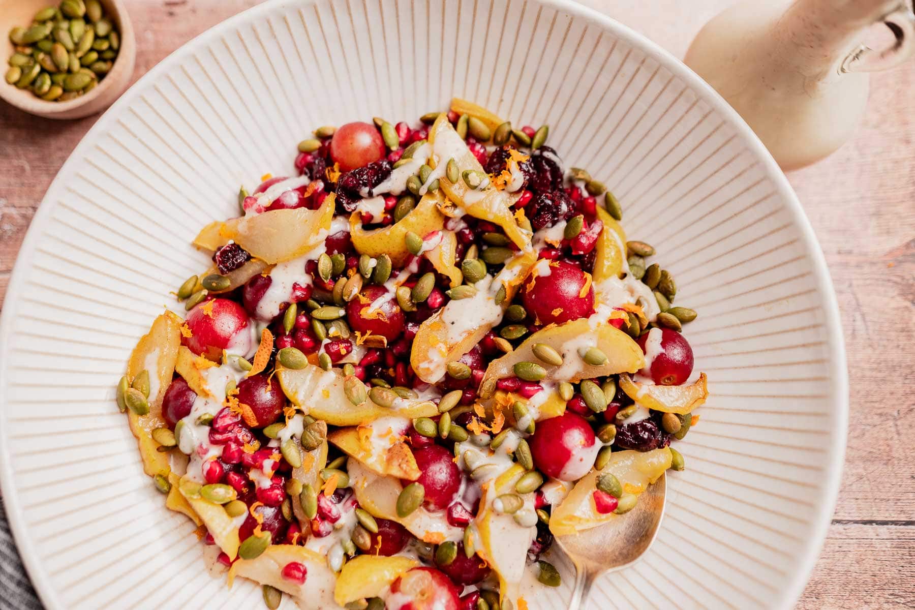 A bowl of Thanksgiving fruit salad with sliced pears, red grapes, pomegranate seeds, dried cranberries, and pumpkin seeds, lightly drizzled with dressing. A spoon rests in the bowl.