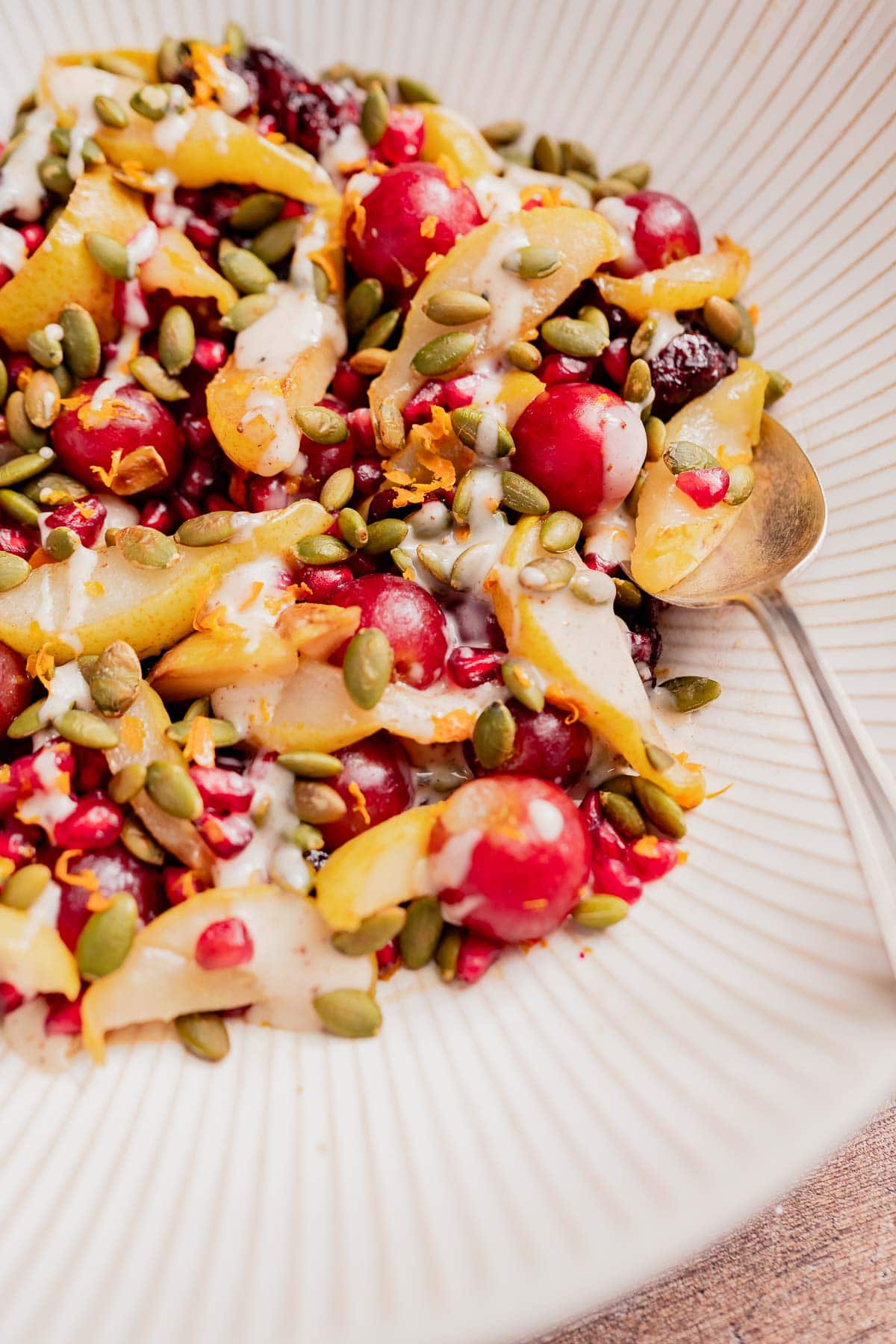 A bowl of thanksgiving fruit salad with pomegranate seeds, sliced pears, cherries, and pumpkin seeds topped with a light creamy dressing, served with a spoon on the side.
