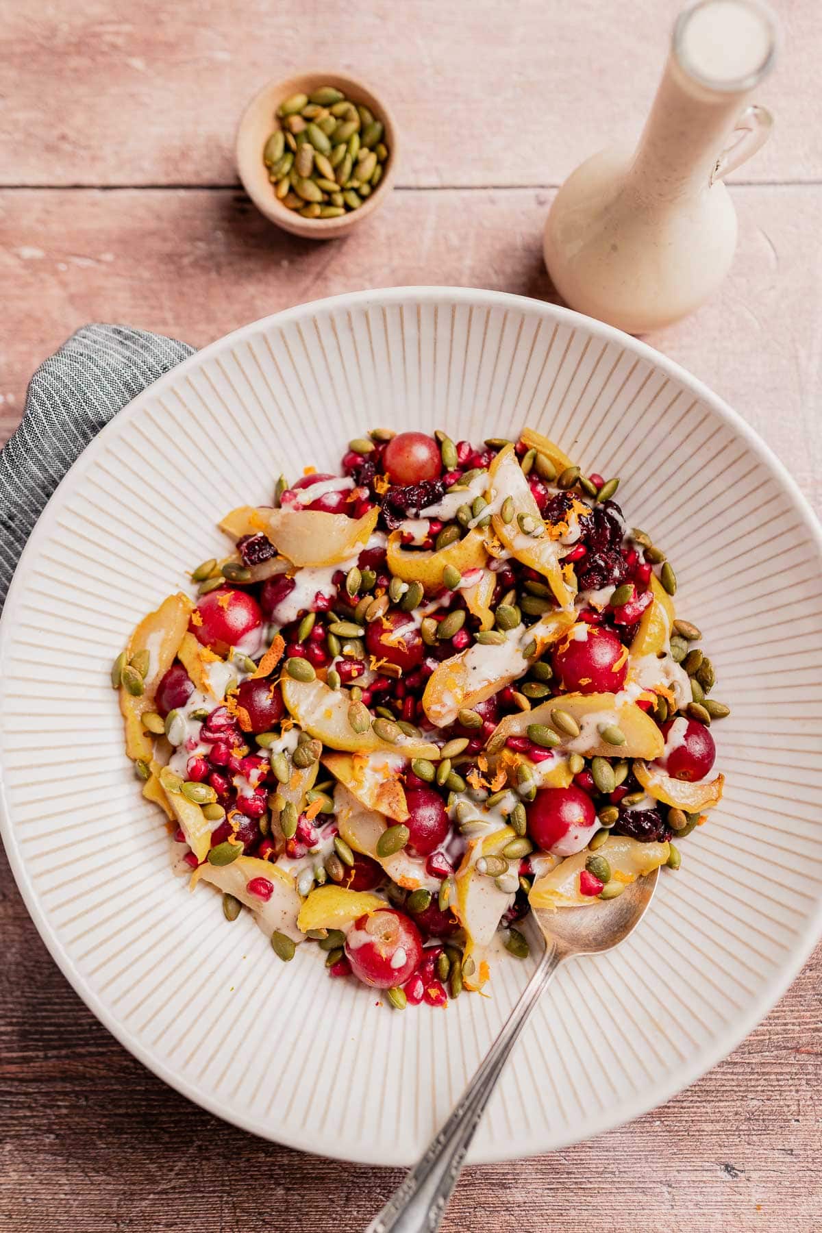 A bowl of thanksgiving fruit salad with sliced pears, red grapes, pomegranate seeds, pumpkin seeds, and dressing sits on a wooden table beside a small bowl of seeds and a bottle of dressing.