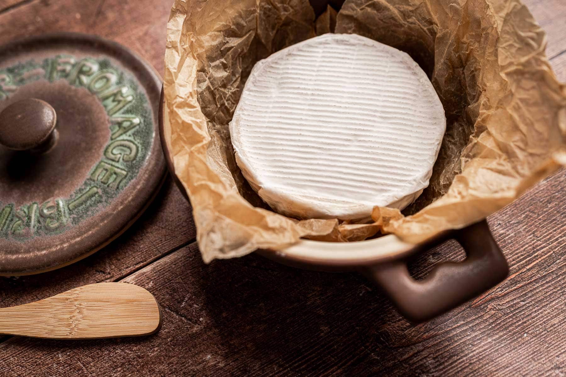 A round of soft white cheese, likely Brie and fig jam, sits on brown parchment in a ceramic dish beside a wooden spreader and a cheese dish lid on a wooden surface.