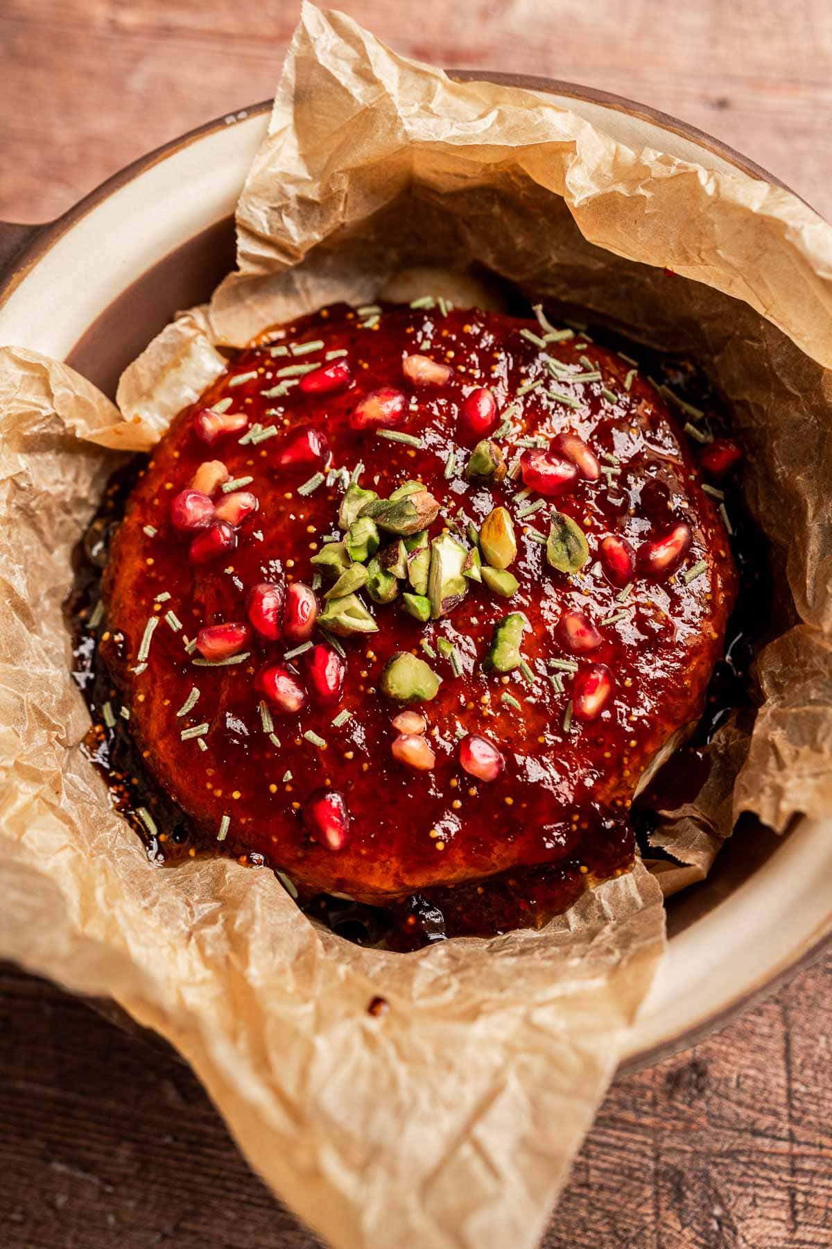 A round baked brie topped with fig jam, pomegranate seeds, herbs, and nuts sits on parchment paper in a dish on a wooden surface.