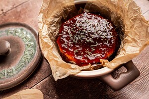 A round baked brie covered in glossy fig jam and sprinkled with herbs sits in parchment paper inside a ceramic baking dish.