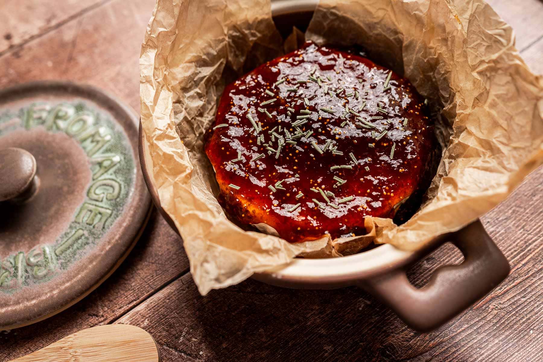 A round baked brie covered in glossy fig jam and sprinkled with herbs sits in parchment paper inside a ceramic baking dish.