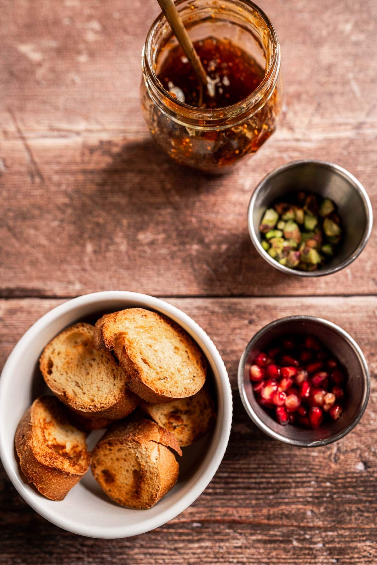 A bowl of toasted bread slices, creamy brie and fig jam, and small bowls of chopped pistachios and pomegranate seeds sit on a wooden surface.
