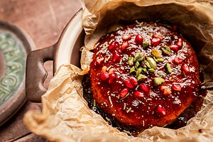 A round baked brie and fig jam cheese, topped with pomegranate seeds, chopped pistachios, and herbs, sits on parchment paper in a ceramic dish.