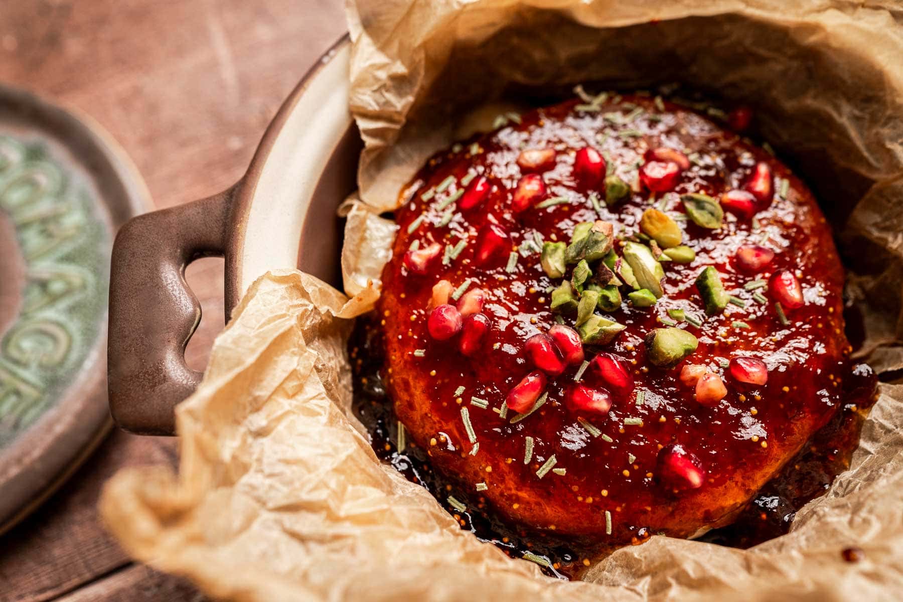 A round baked brie and fig jam cheese, topped with pomegranate seeds, chopped pistachios, and herbs, sits on parchment paper in a ceramic dish.