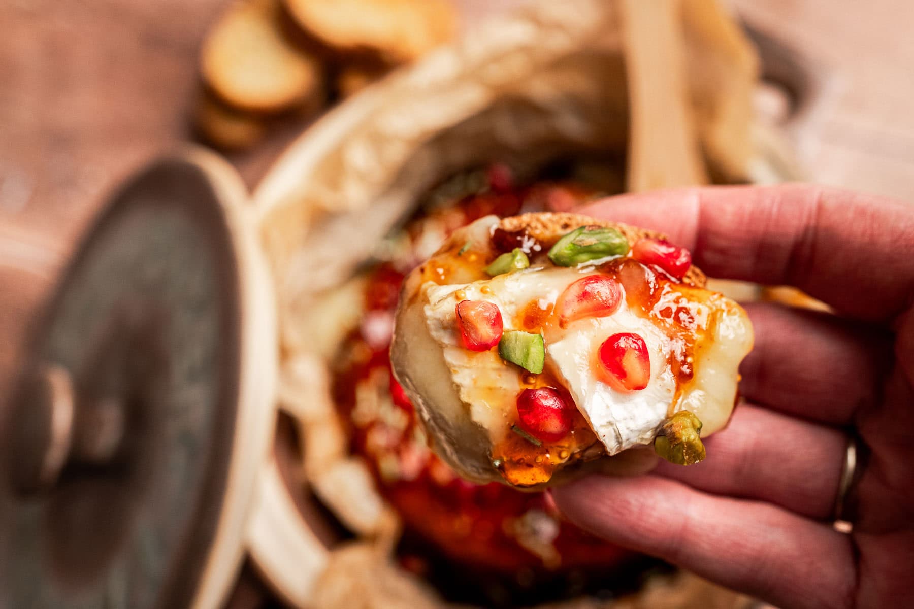 A hand holds a piece of bread topped with brie and fig jam, melted cheese, pomegranate seeds, green garnish, and sauce, with more bread and cheese visible in the background.