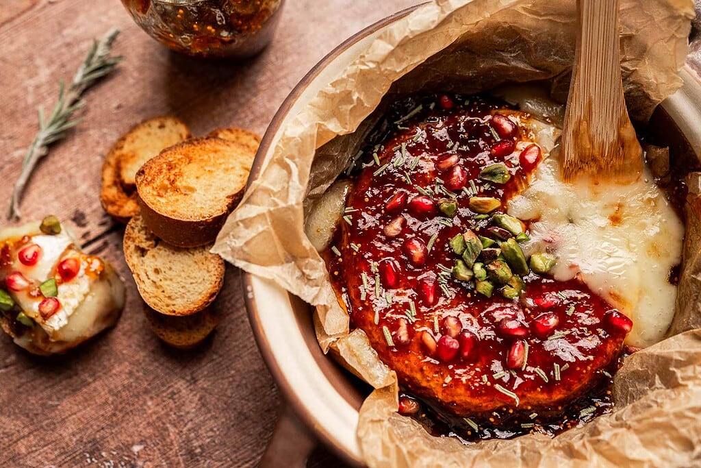 Round baked brie and fig jam cheese topped with pomegranate seeds, pistachios, and herbs, partially sliced in brown parchment paper, served with toasted bread slices and a jar of preserves nearby.