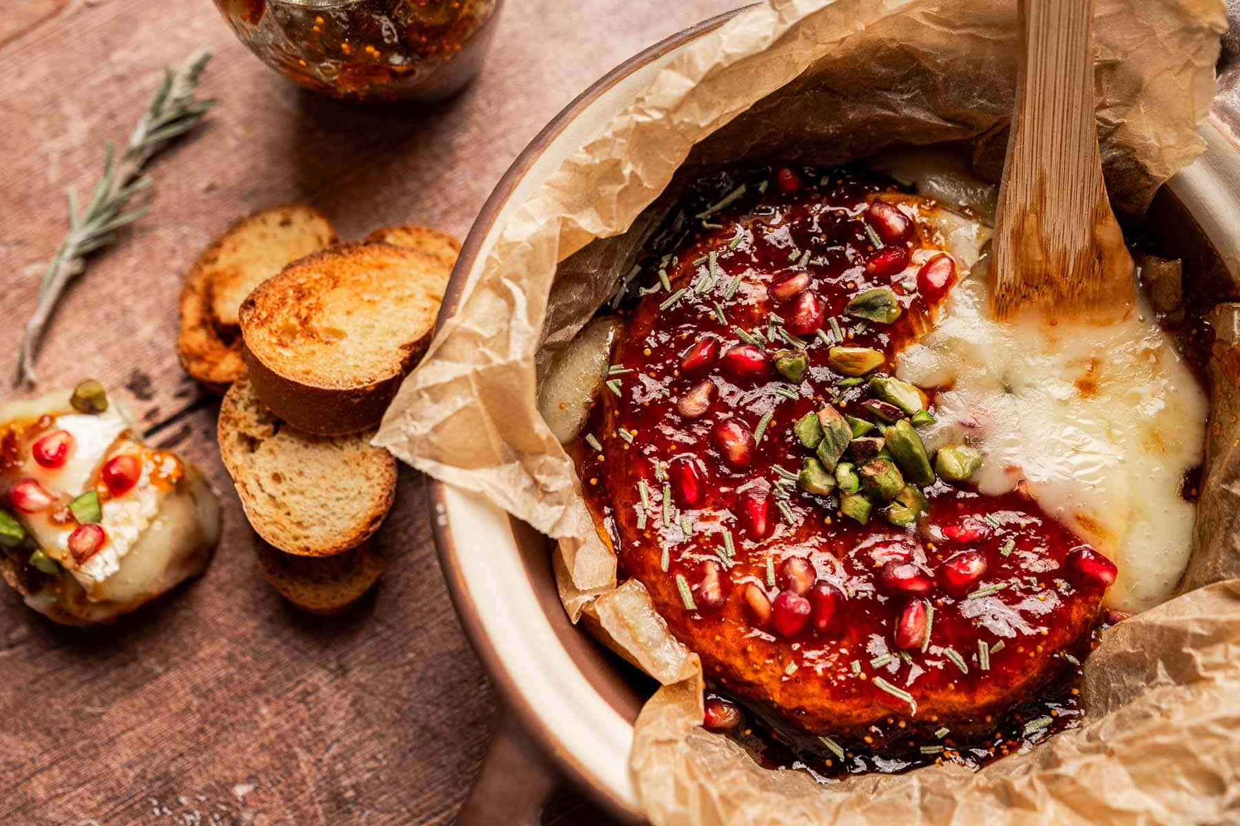 Round baked brie and fig jam cheese topped with pomegranate seeds, pistachios, and herbs, partially sliced in brown parchment paper, served with toasted bread slices and a jar of preserves nearby.